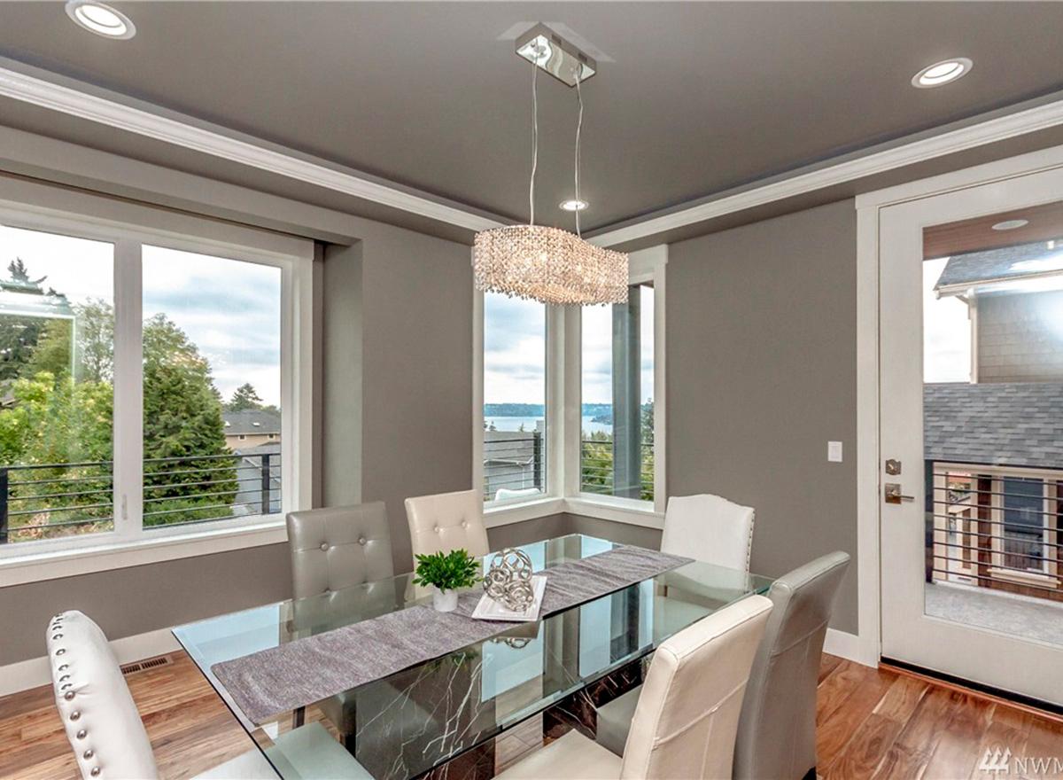 Dining room with glass table, upholstered chairs, large windows, and crystal chandelier.