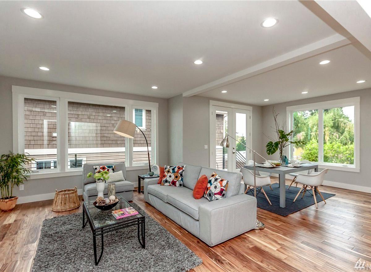 Open concept living and dining areas featuring a gray sofa, glass-top coffee table, and dining set with chairs by large windows.