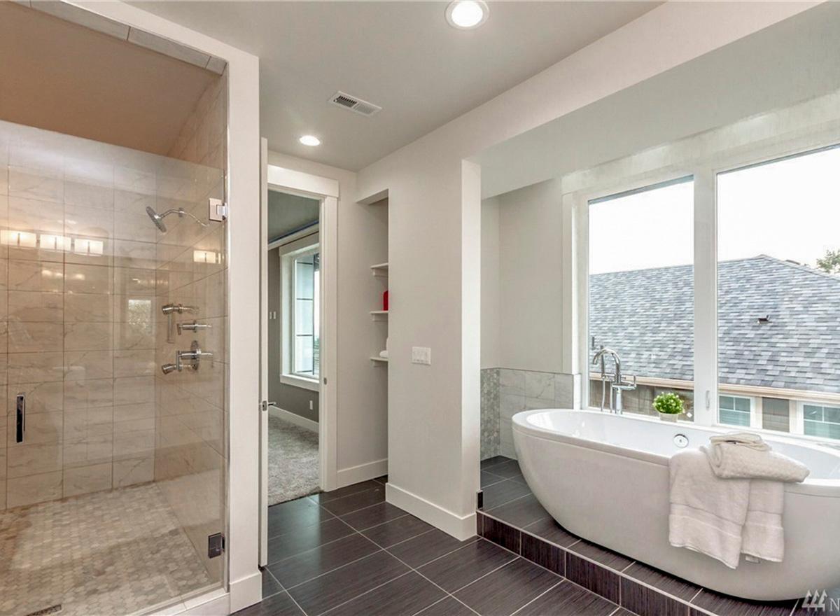Modern bathroom with freestanding tub, walk-in shower with marble tile, and large windows overlooking rooflines.