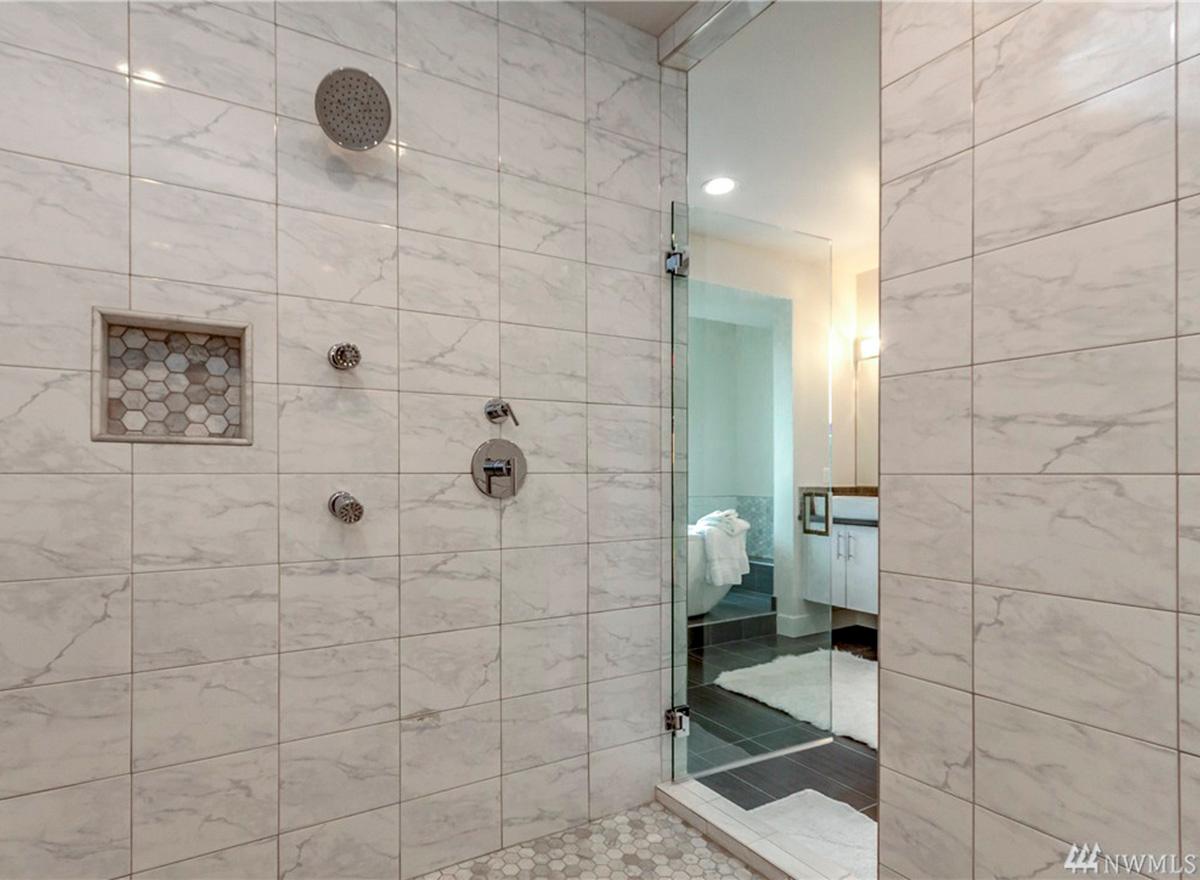 Bathroom interior with marble tile shower, multiple shower heads, glass enclosure, and visible bathtub and vanity.