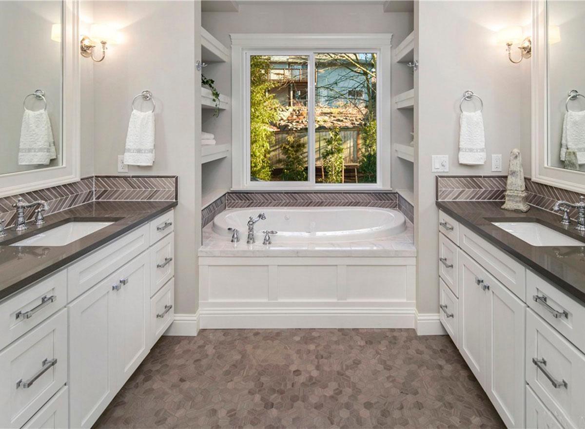 Bathroom with dual vanities, soaking tub centered under a large window, herringbone tile backsplash, and hexagonal floor tiles.