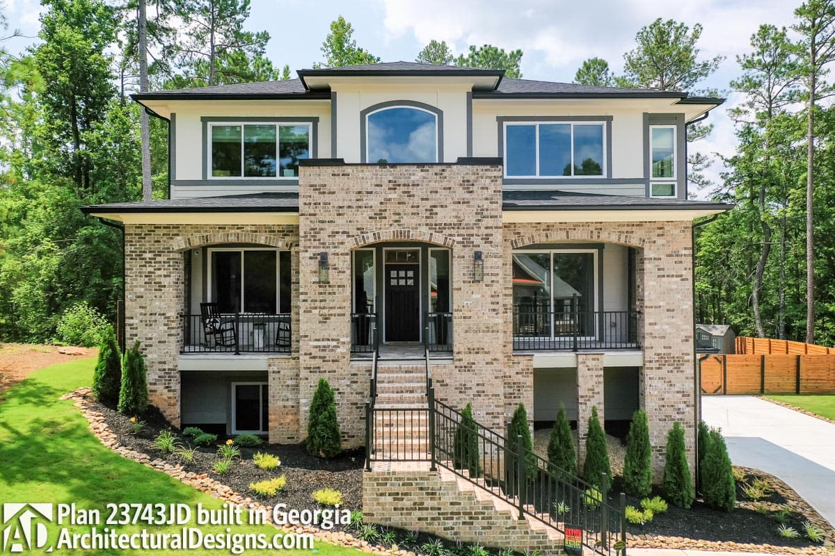 House plan exterior: two-story home with brick facade, arched entry, second-story balcony, and large windows.