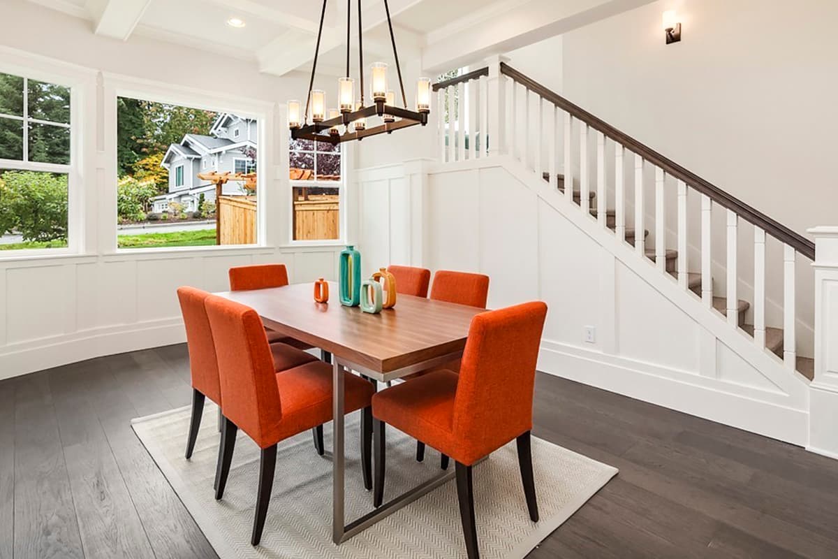Dining room with wood table, orange chairs, coffered ceiling, and staircase. Large windows offer exterior view.