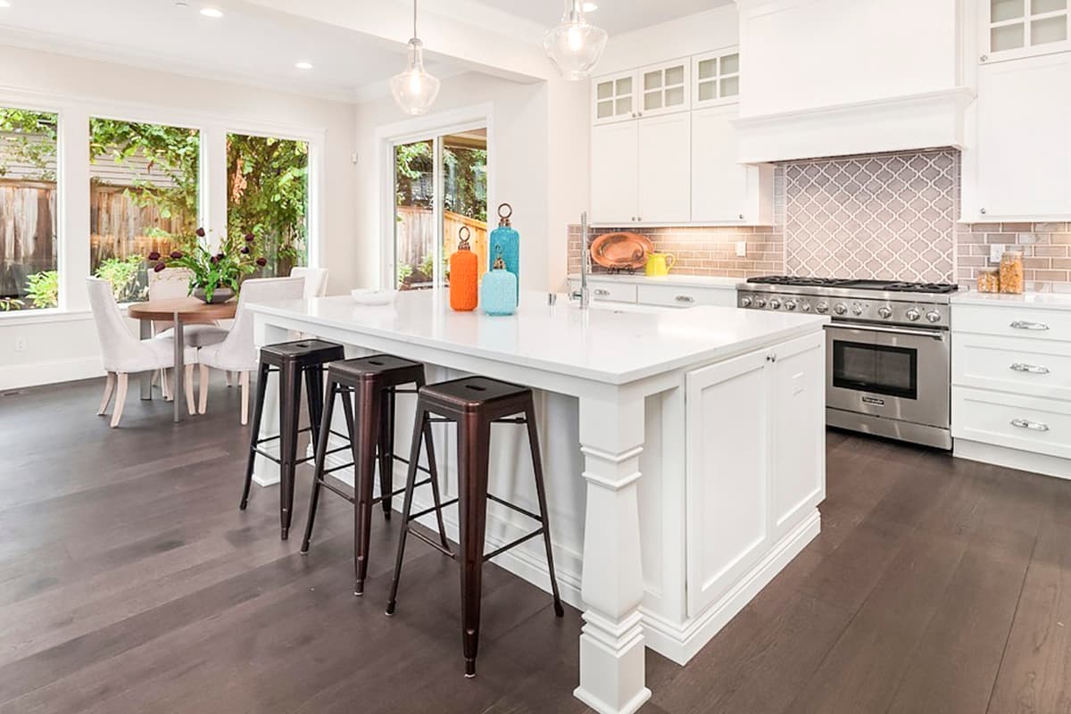 Kitchen with white island, bar stools, stainless steel range, white cabinetry, pendant lights, and large windows overlooking the yard.