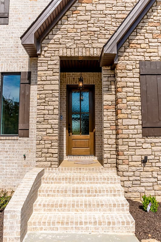 Exterior house plan: Brick and stone facade with wooden door, covered entryway, gabled dormers, and shuttered windows.