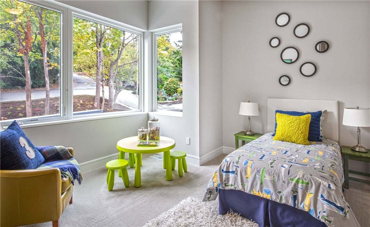 Children's bedroom with corner windows, a small green table and stools, a yellow armchair, and a twin bed with airplane-themed bedding.