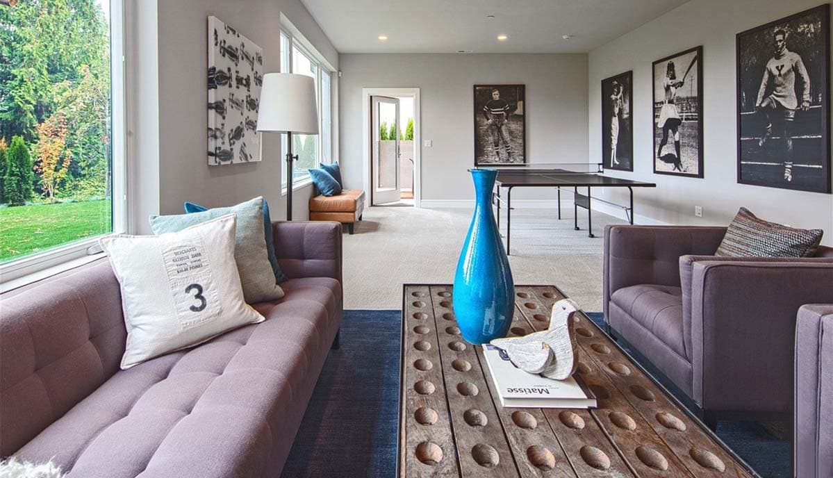Living room with large windows, tufted sofa, armchairs, coffee table with blue vase, and ping pong table.