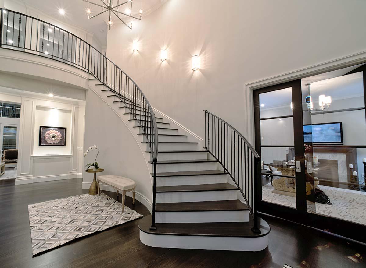 Interior view of a grand foyer with a curved staircase featuring black railings, dark wood treads, and white risers. A balcony overlooks the space.