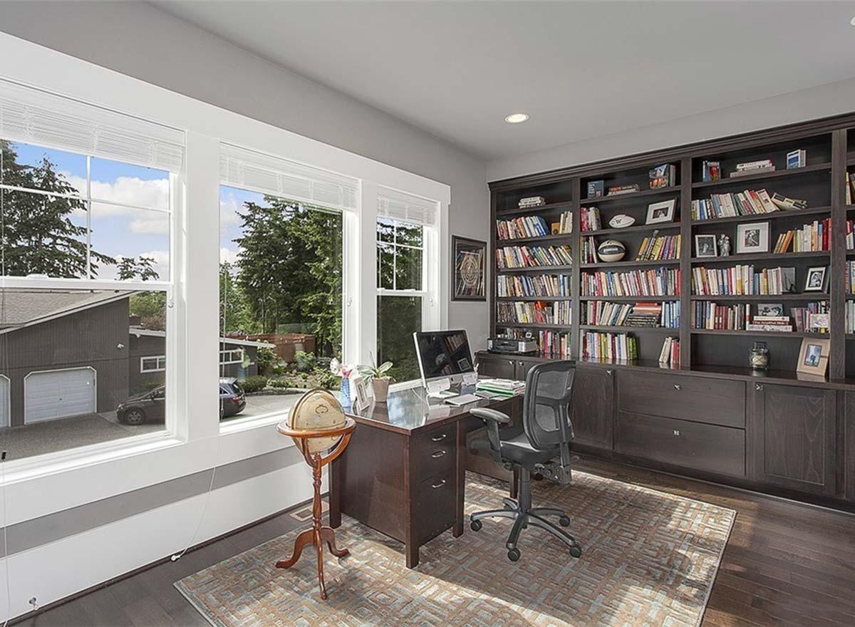 Home office with large built-in bookshelves, a desk with computer, and large windows overlooking a garage and trees.