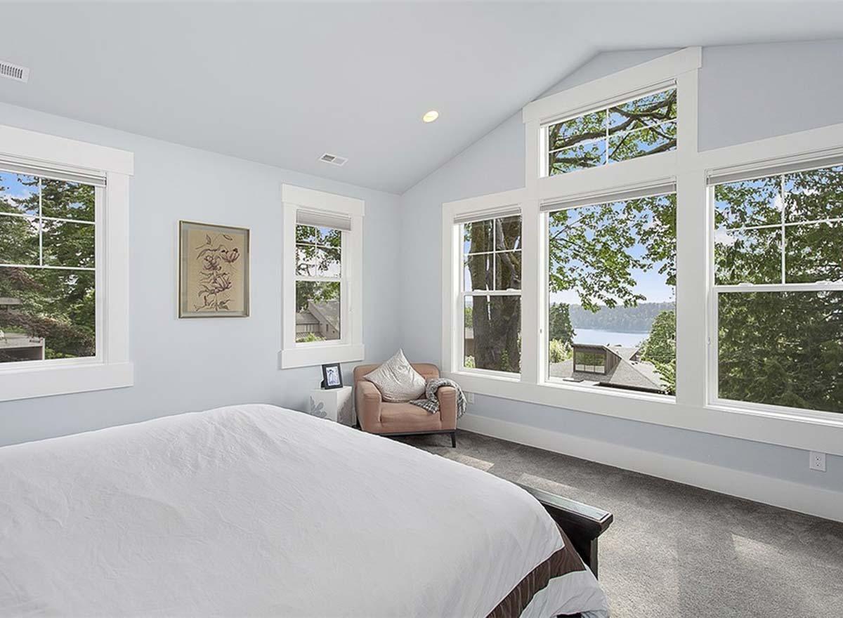 Bedroom interior with vaulted ceiling, large grid windows showcasing trees and a body of water, and a single chair.