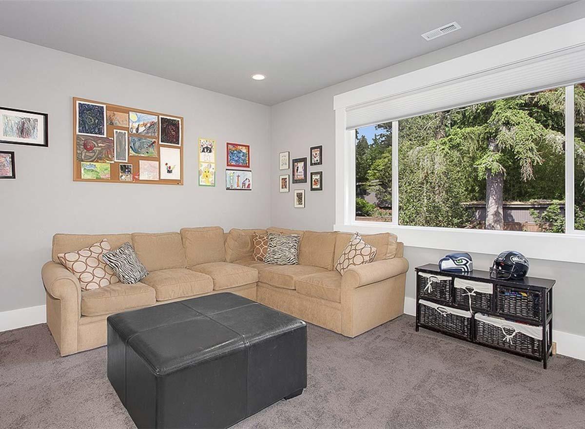 Living room with tan sectional sofa, black ottoman, corkboard art display, and large window overlooking trees.