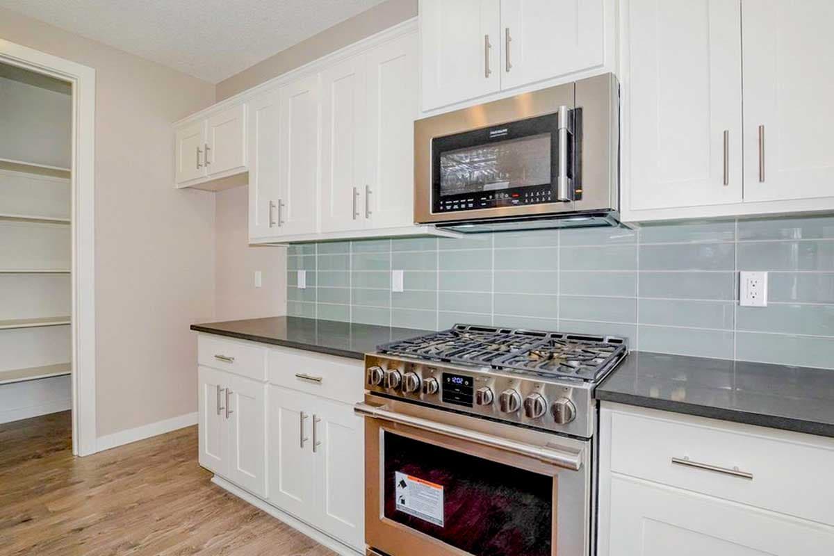 Kitchen interior with white cabinetry, stainless steel gas range, microwave, and pale blue subway tile backsplash. Adjacent pantry visible.