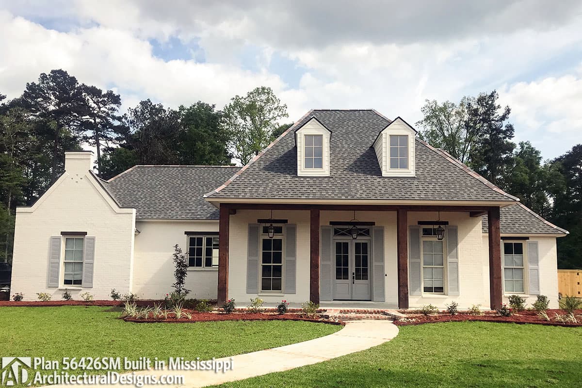 House plan exterior: One-story home with central covered porch supported by timber posts, two dormer windows, and a brick chimney.