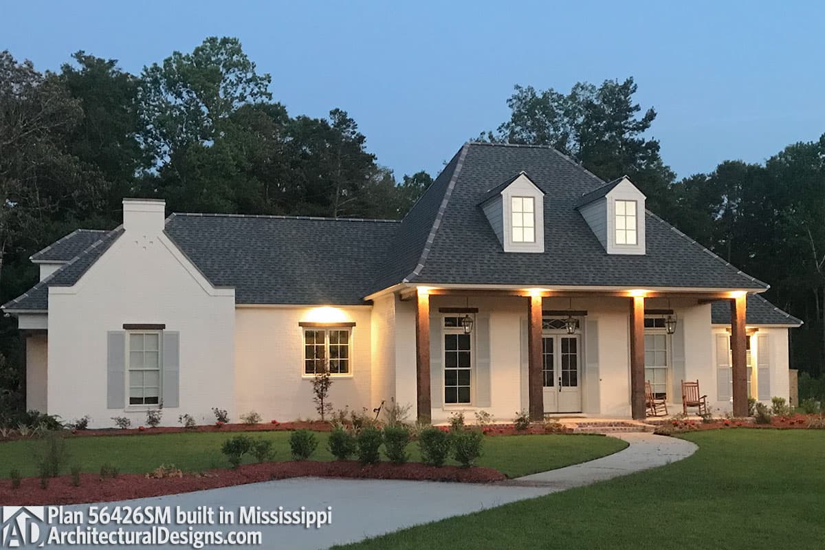 House plan exterior. One-story white brick home with a steeply pitched roof, dormers, and a covered porch with wood beams.