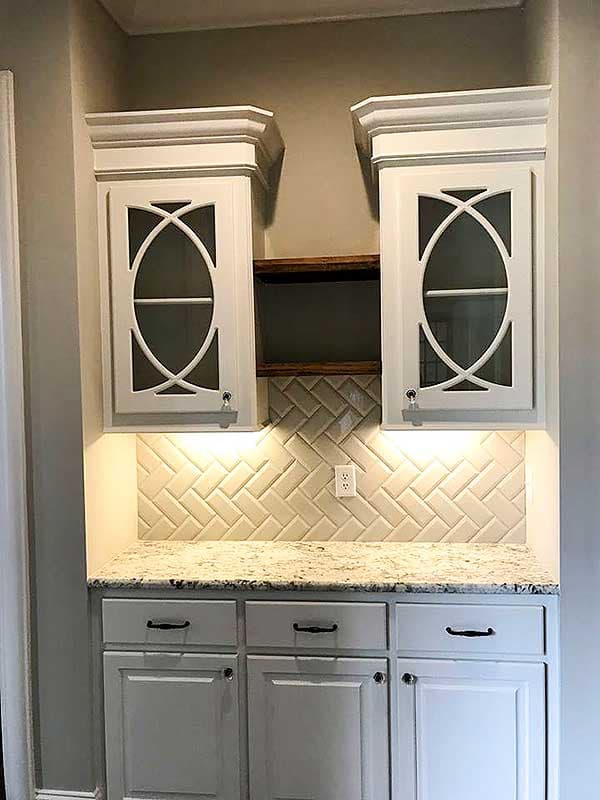 Interior view of a kitchen bar area with white cabinets, granite countertop, herringbone tile backsplash, and decorative glass cabinet doors.