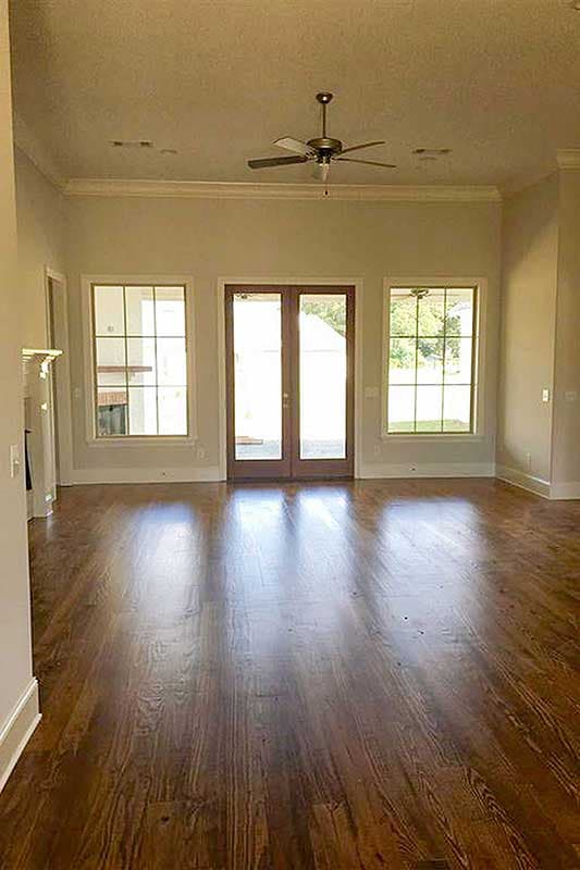 Interior view of a great room with hardwood floors, a ceiling fan, and French doors flanked by two large windows.