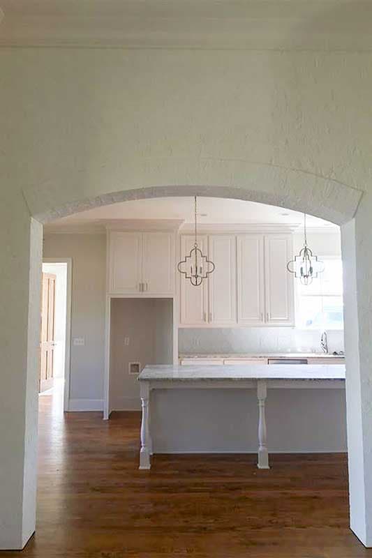 Interior view of a kitchen featuring white cabinets, a marble-topped island, and decorative pendant lights, seen through an arched doorway.