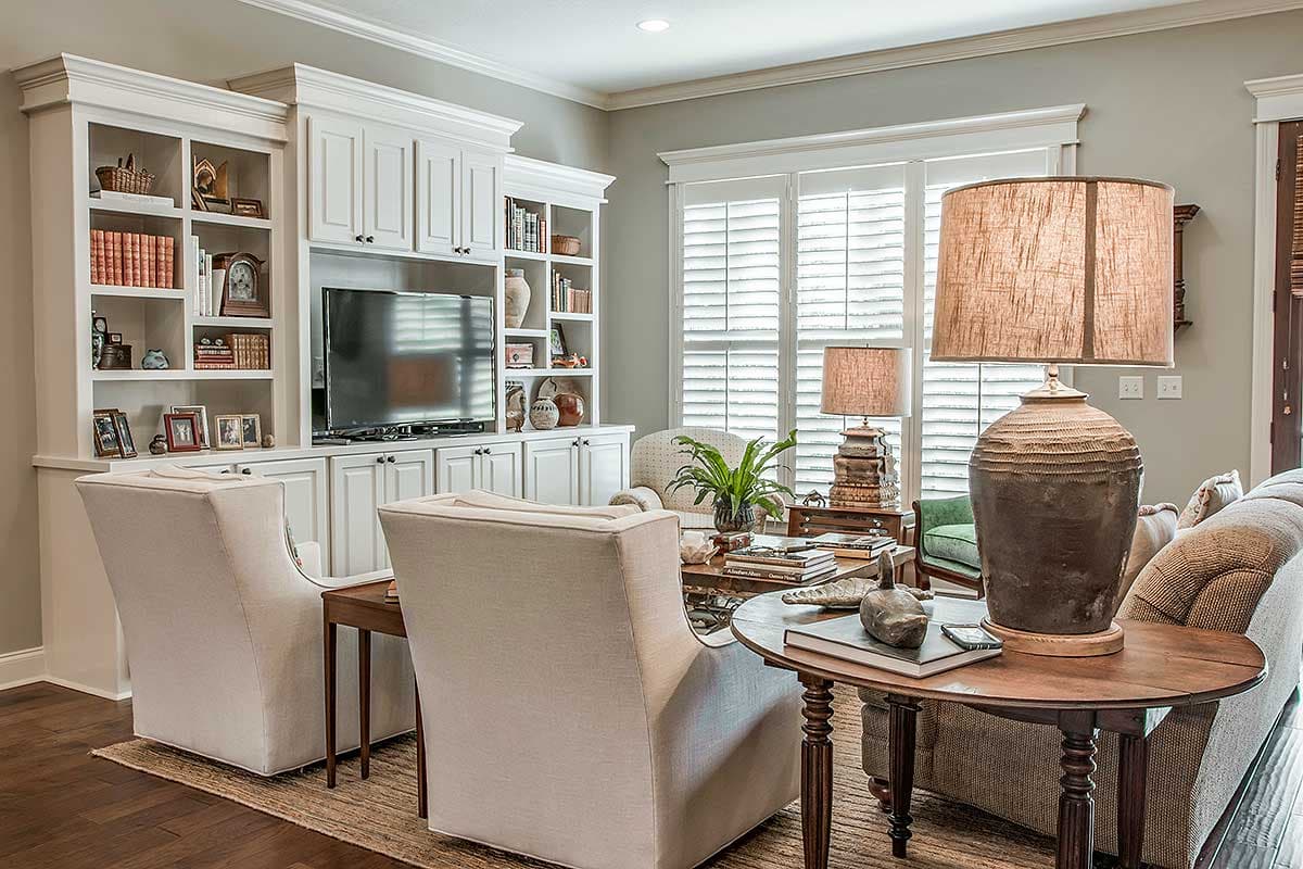 Living room with built-in white cabinetry, TV, and plantation shutters. Includes two armchairs, lamps, and decorative objects.