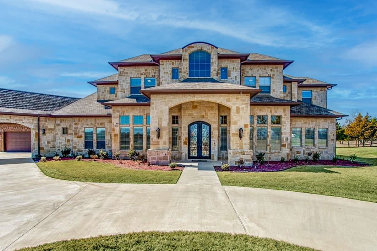 House plan exterior. Two-story with stone facade, arched entry, multiple gables, and porte-cochere garage with tile roof. Large windows throughout.
