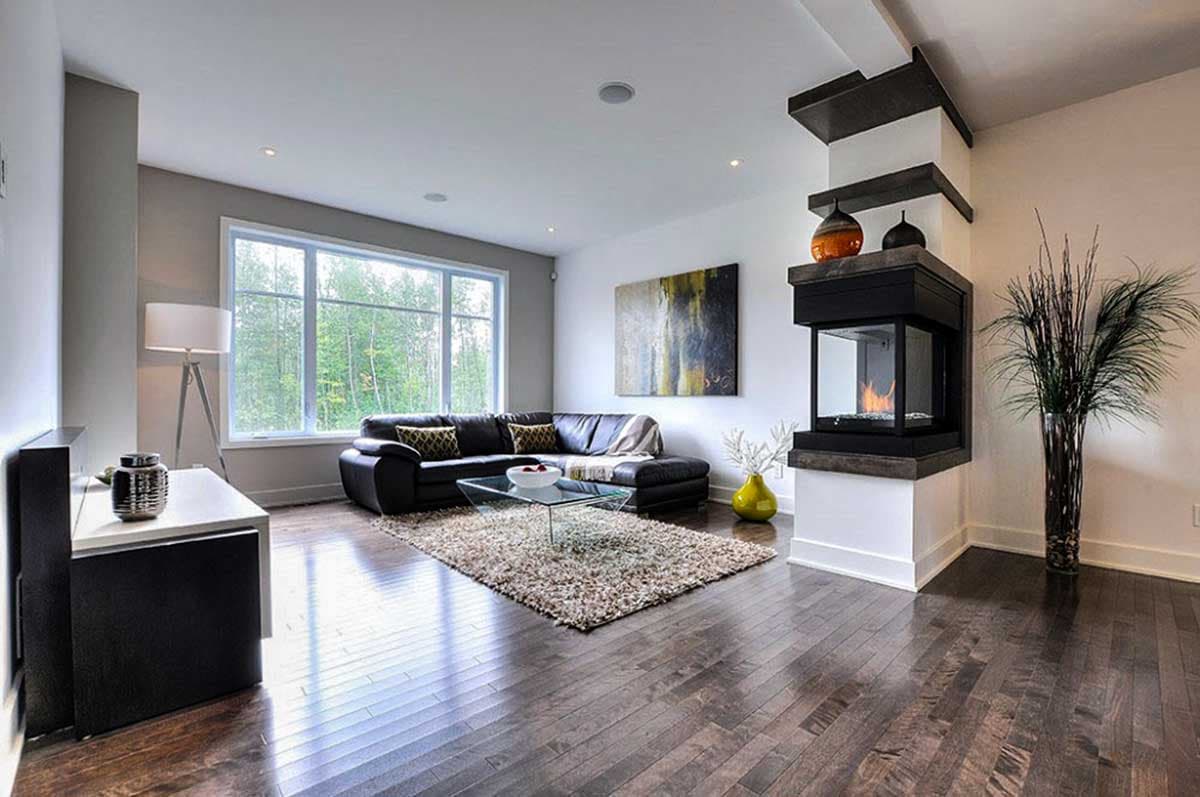 Modern living room with black leather sofa, glass coffee table, fireplace, and large window overlooking trees.