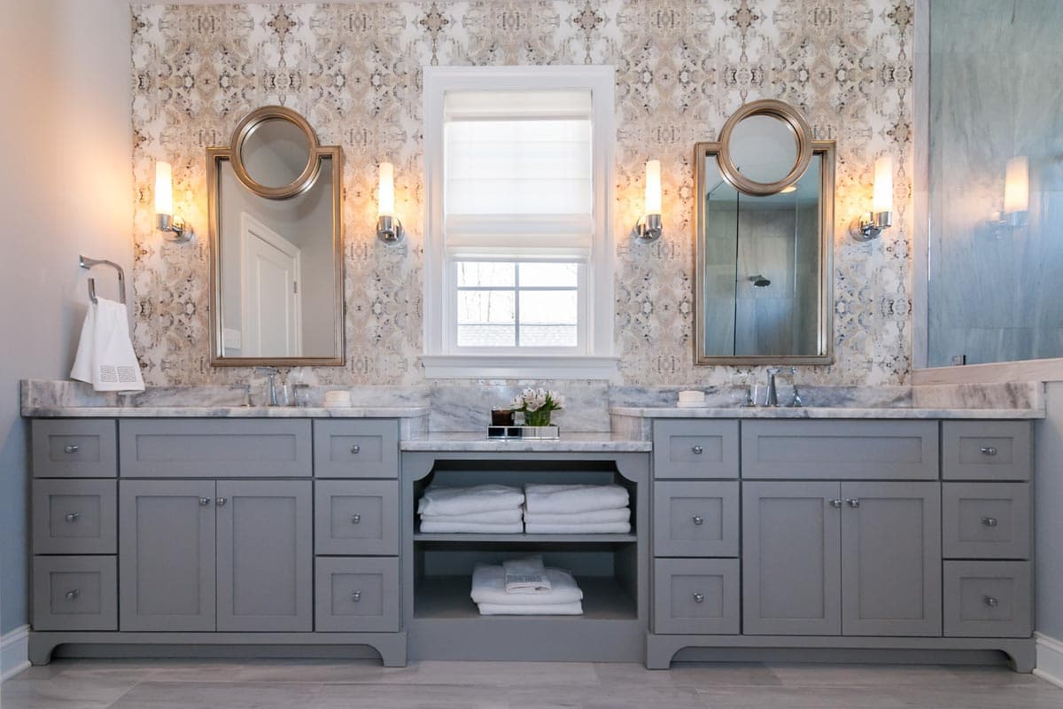 Bathroom with dual gray vanities, marble countertops, mirrored cabinets, ornate wall sconces, and patterned wallpaper.