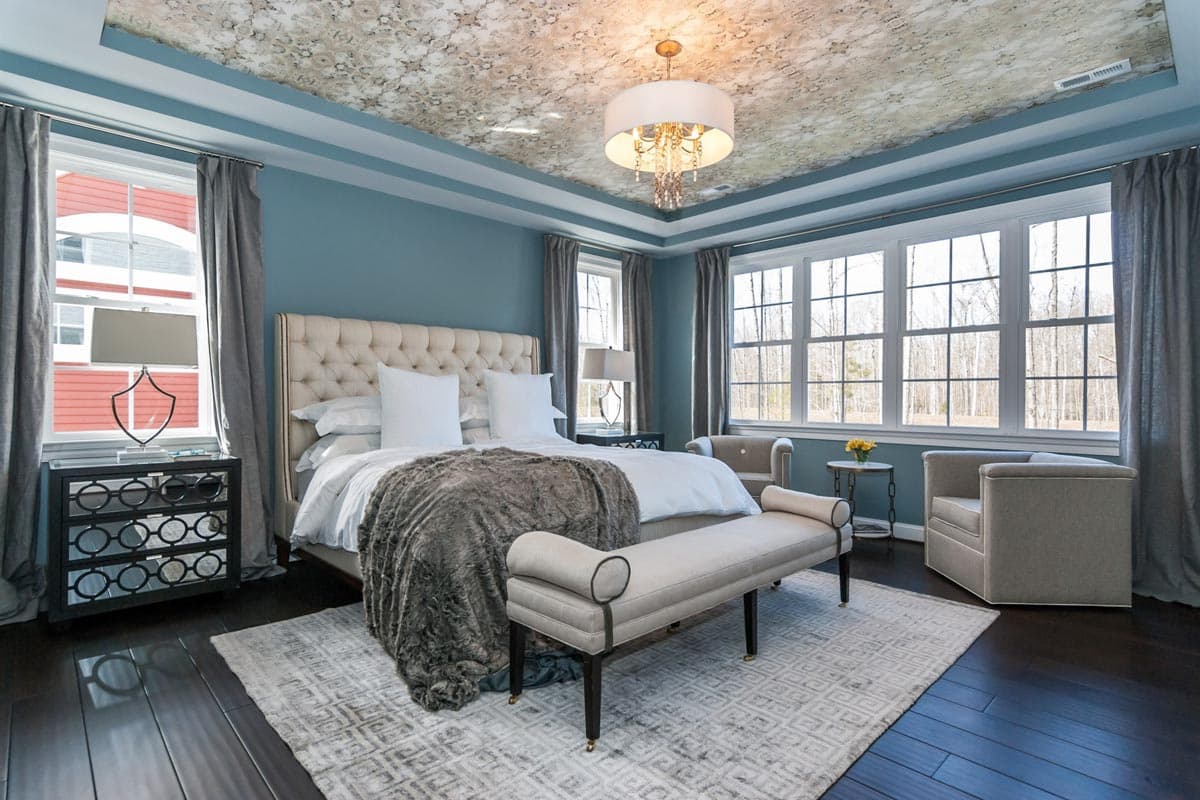 Bedroom with tufted headboard, patterned ceiling, large windows overlooking woods, and seating bench.