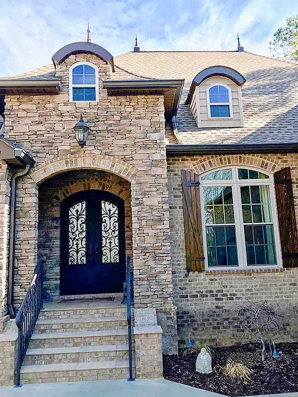 House plan exterior with stone and brick facade, arched entryway, wrought iron double doors, and arched dormer window.