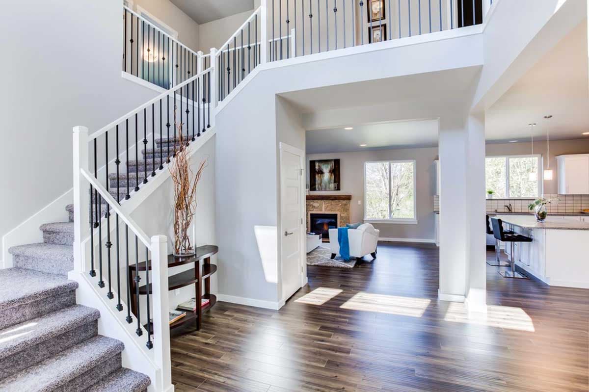 Interior view of a two-story foyer with a carpeted staircase, black spindles, and view into the living room and kitchen.