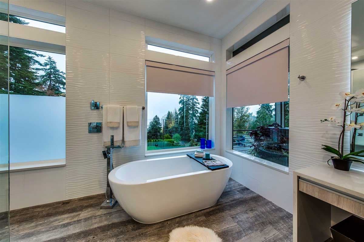 Modern bathroom with freestanding tub, large windows, textured tile wall, and wood-look flooring.