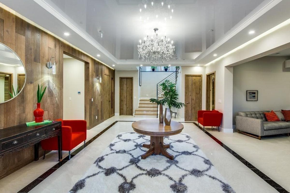 Interior view of a modern foyer with wood-paneled walls, a chandelier, stairs, and a round dining table on a patterned rug.