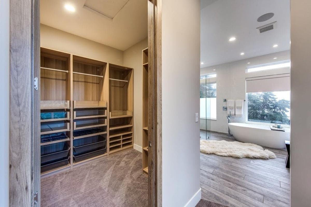 Walk-in closet with wood shelving and drawers next to a modern bathroom with a freestanding tub and large windows.