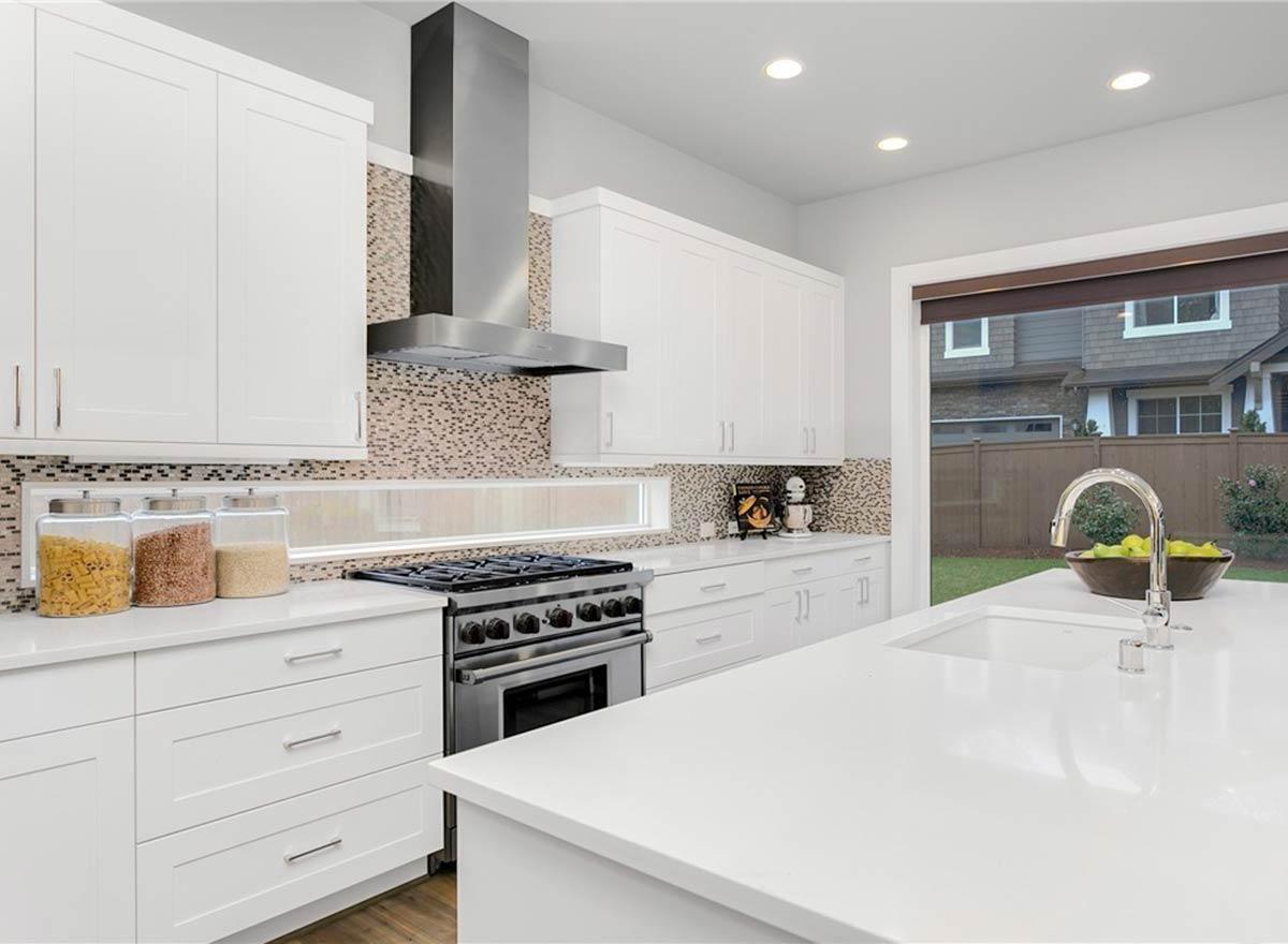 Modern kitchen with white cabinets, stainless steel range hood, mosaic tile backsplash, and island with sink. Outside view shows backyard.
