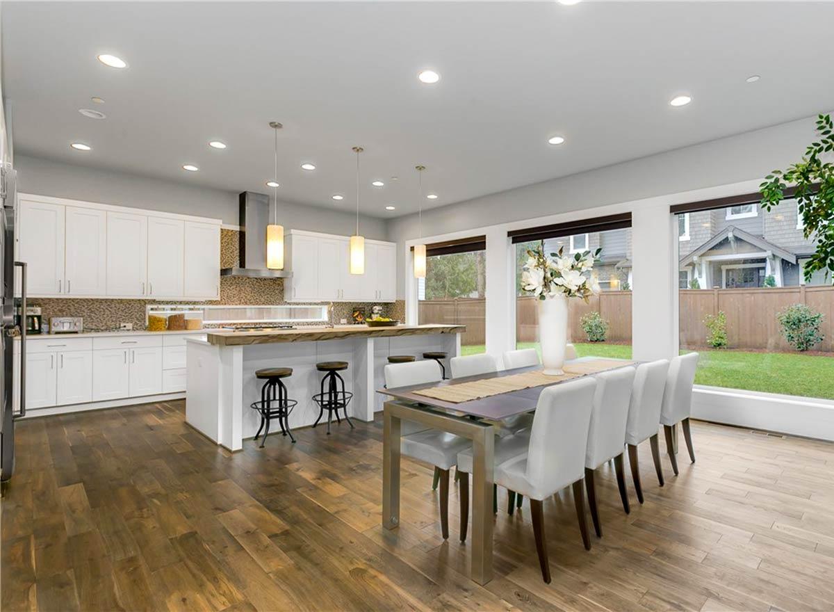 Open-concept kitchen and dining area with white cabinets, island, and large windows overlooking a backyard.
