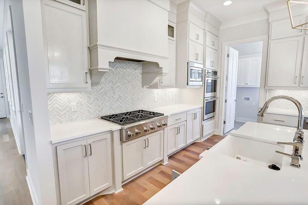 Kitchen interior with white cabinets, gas range, double ovens, herringbone tile backsplash, and a farmhouse sink with gooseneck faucet.