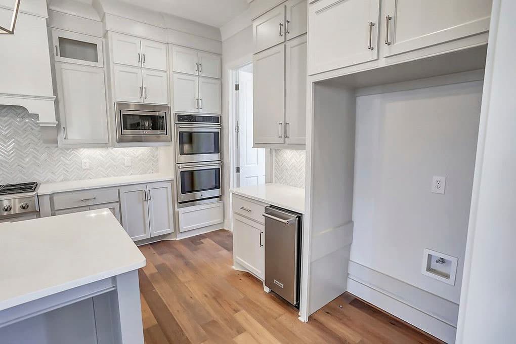 Modern kitchen interior with white cabinetry, double ovens, herringbone tile backsplash, island, and hardwood floors.