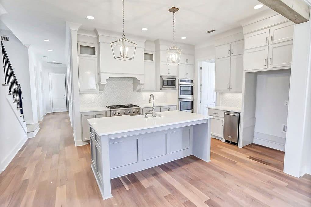 Kitchen with island, gas range, double ovens, herringbone backsplash, and white cabinets.