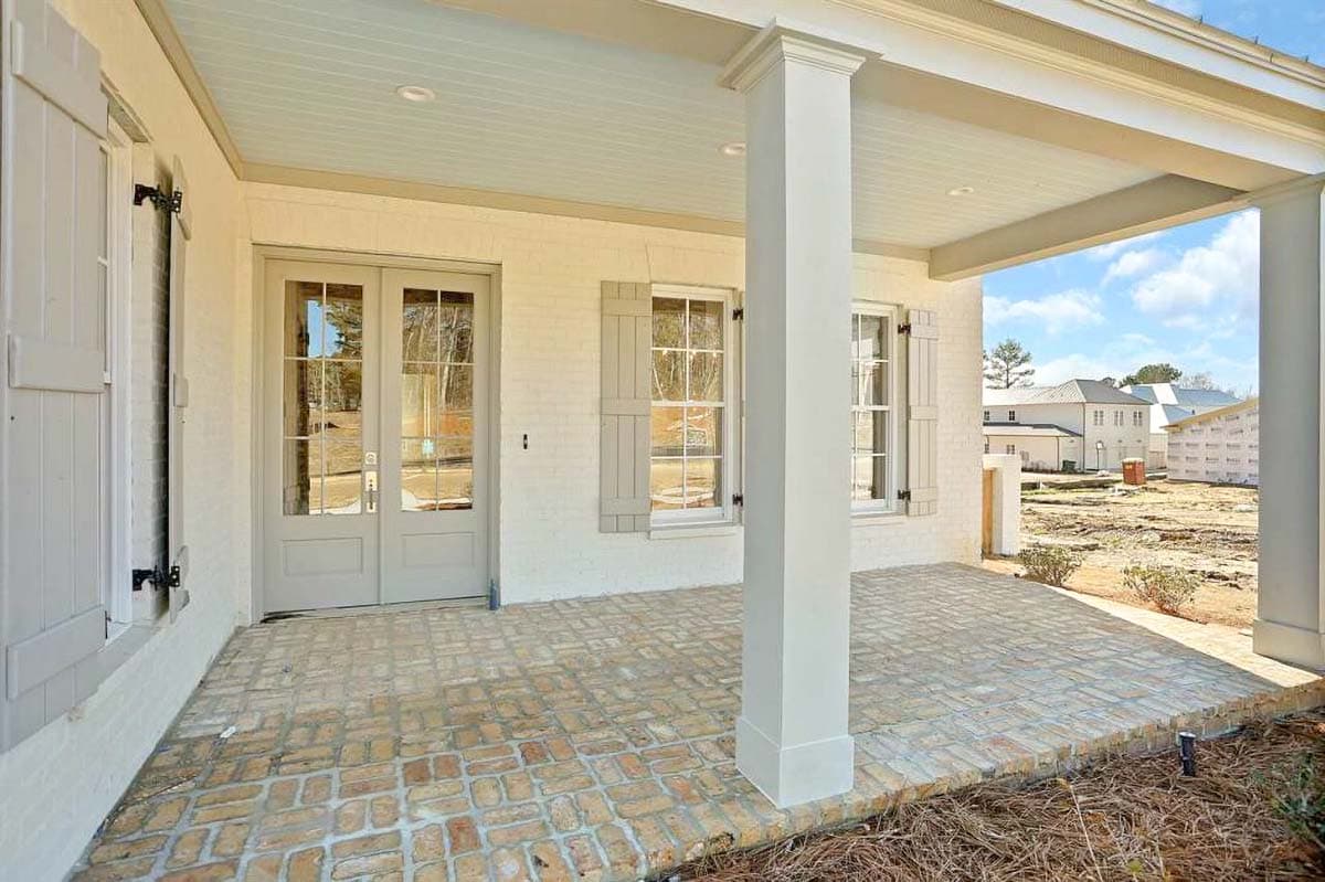 House plan exterior with white brick, double glass doors, paneled shutters, and covered porch with brick paver flooring.