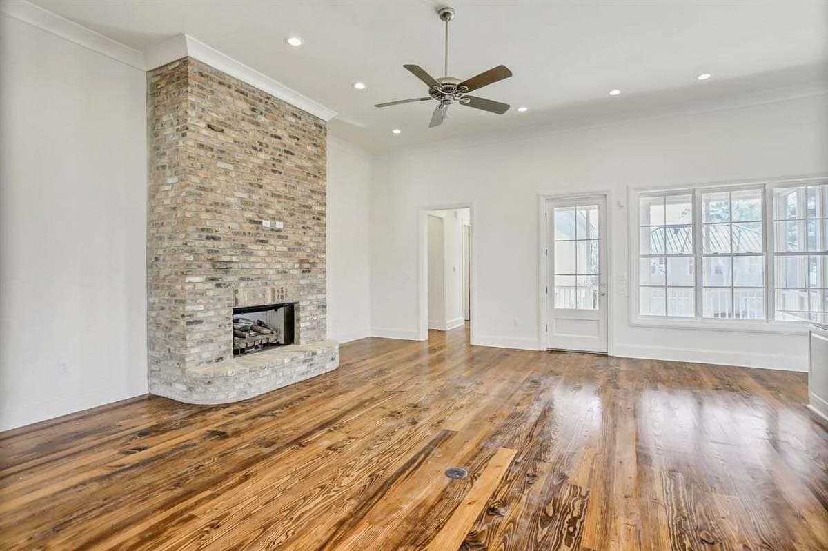 Living room interior with rustic brick fireplace, hardwood floors, ceiling fan, and large windows.