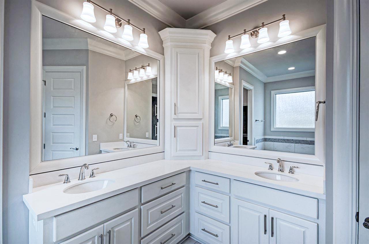 Bathroom vanity with double sinks, white countertops, and a corner linen cabinet. Overhead lights reflect in large mirrors.