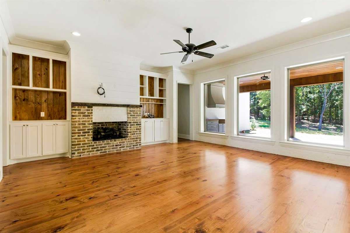 Great room with brick fireplace, built-in shelving, ceiling fan, and large windows overlooking outdoor space.