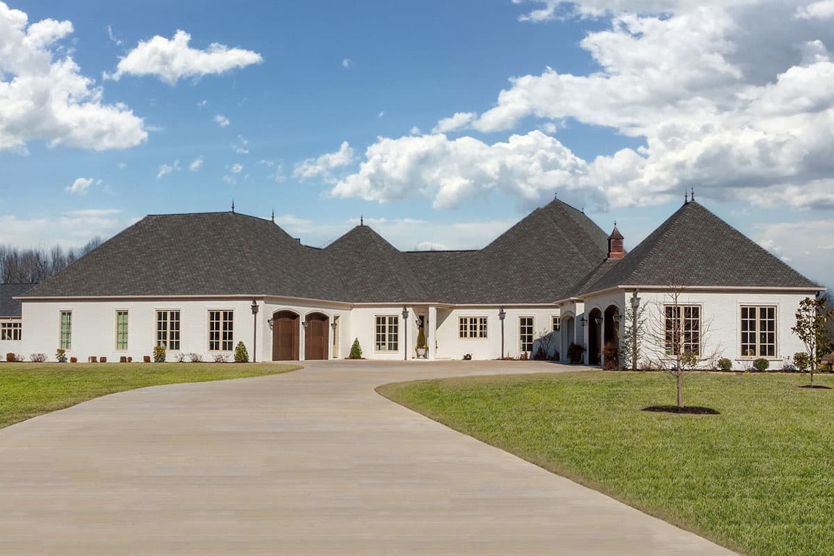 House plan exterior of a sprawling one-story residence with a dark shingled hip roof, white brick facade, and a U-shaped driveway.