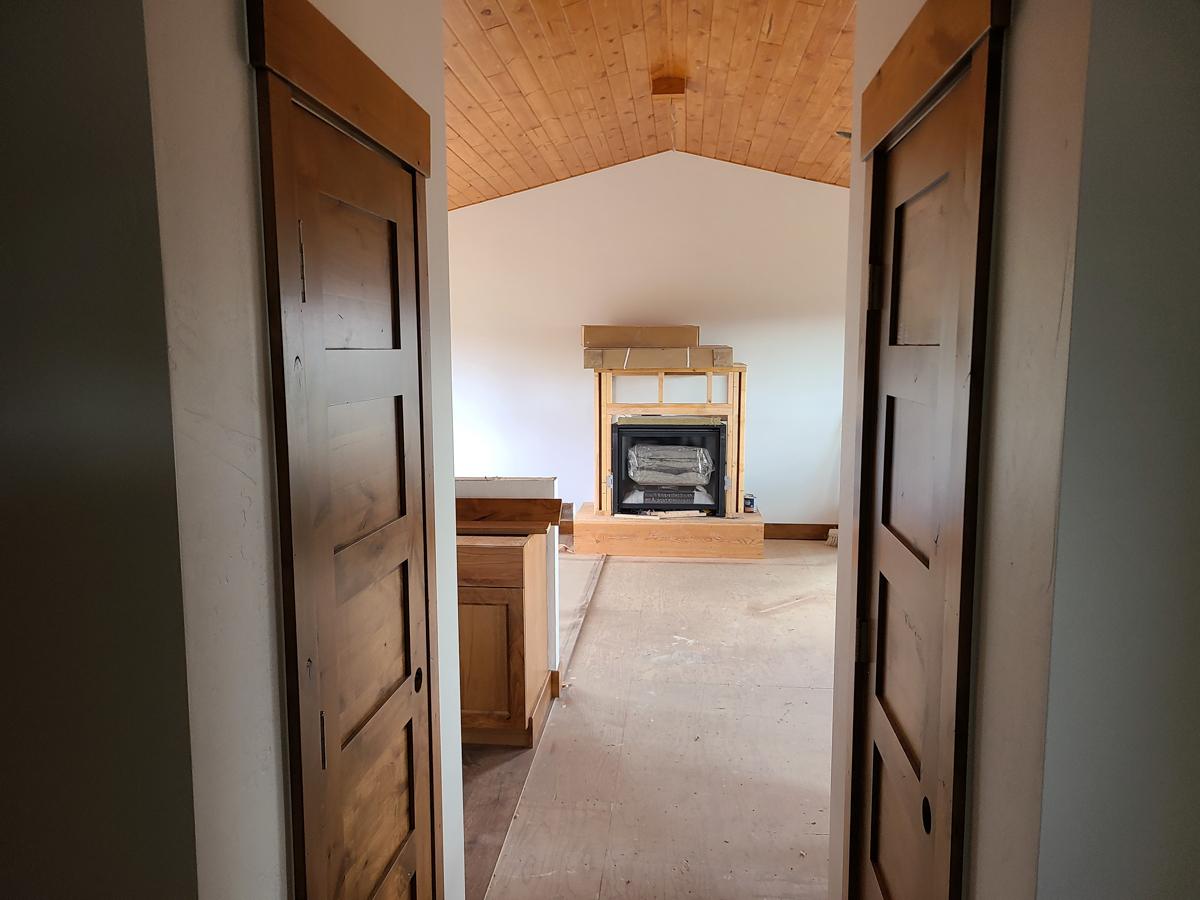 Interior view looking into a room with a fireplace under construction, wood-paneled vaulted ceiling, and wooden sliding doors.