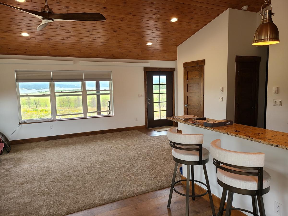 Interior view of a living area with wood-paneled ceiling, large windows, carpeted floor, and a granite countertop bar with stools.