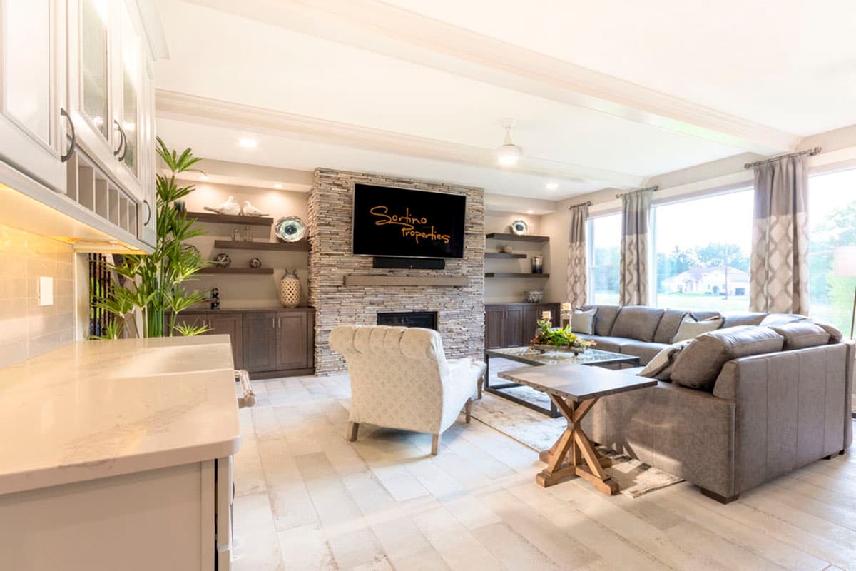 Interior rendering of a great room with stone fireplace, entertainment center, sectional sofa, and a breakfast bar with light wood flooring.