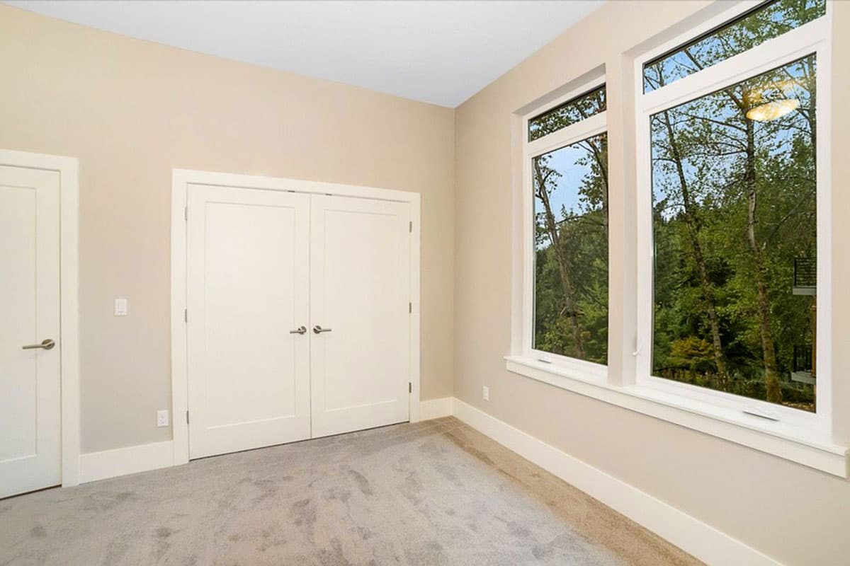 Interior view of room with neutral walls, carpet, double closet doors, and two tall windows overlooking trees.