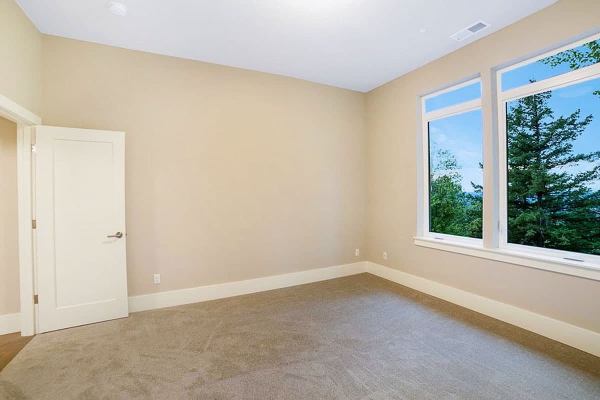 Interior view of an empty room with a closed door, neutral carpet, and large double-pane windows overlooking trees.