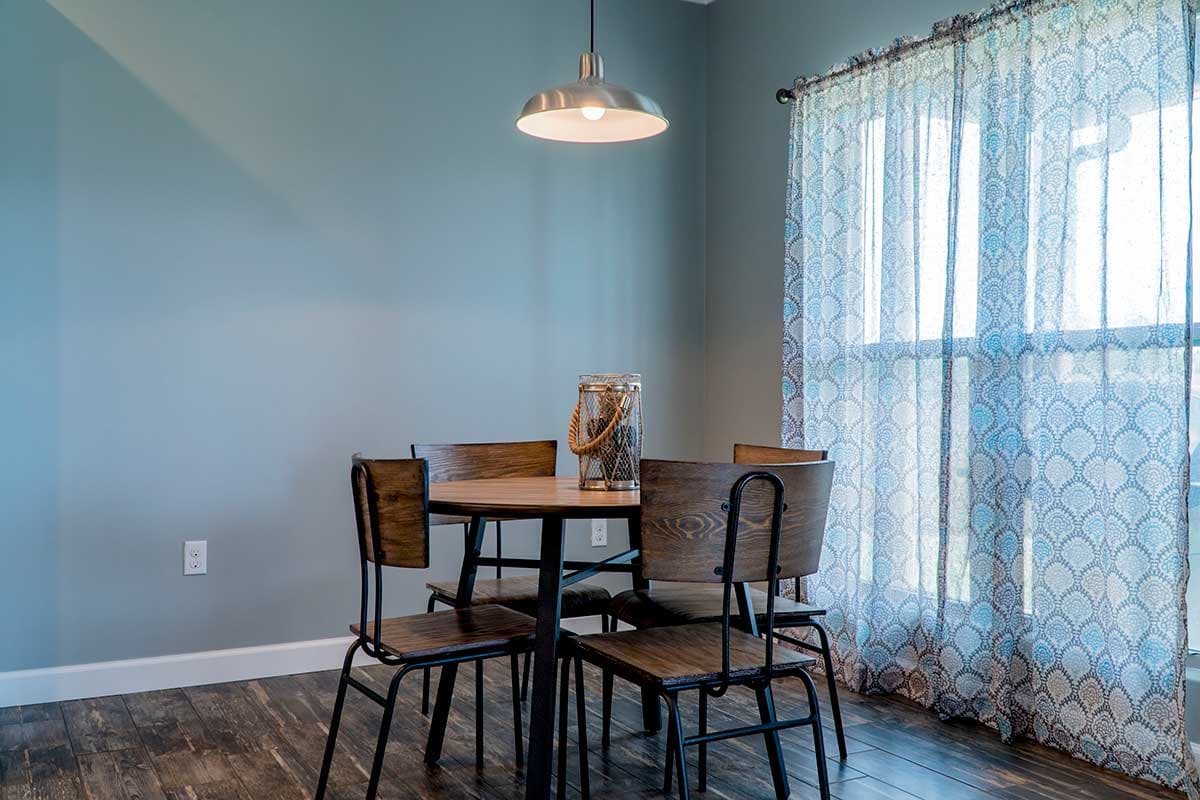 Dining area with round wood table, four chairs, pendant light, and patterned sheer curtains.