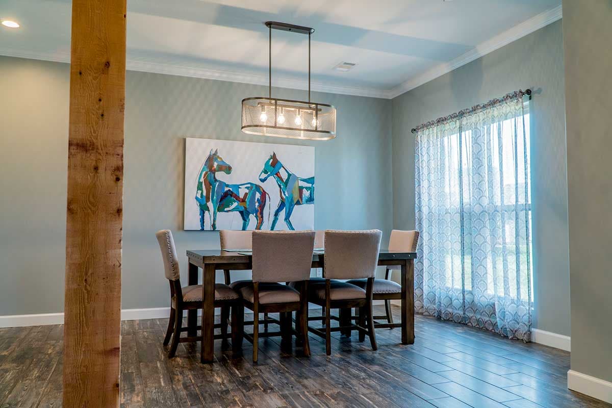 Dining room with dark wood table, upholstered chairs, art featuring horses, and a large window with decorative sheer curtains.