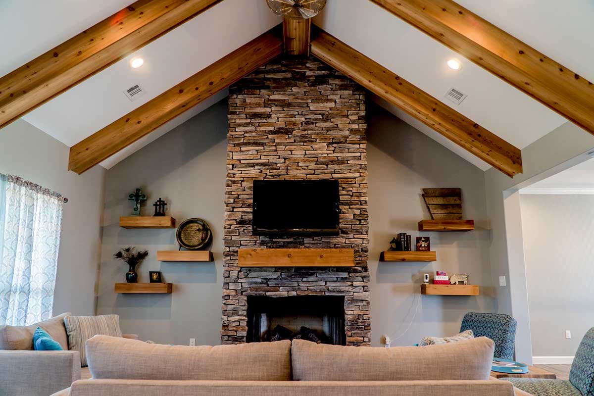 Great room with vaulted ceiling, exposed wood beams, and stone fireplace with mounted television. Includes open shelving.