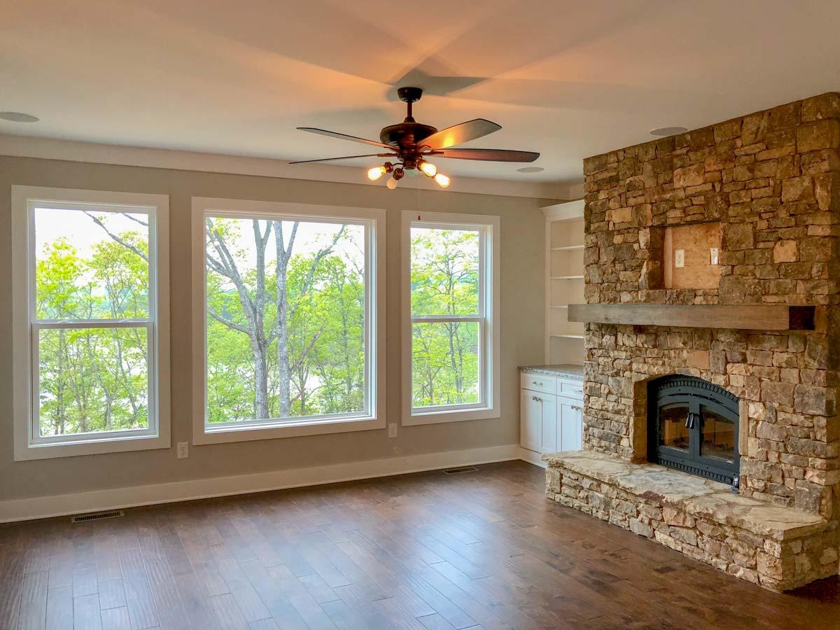 Interior view of a great room with three large windows, a stone fireplace, built-in bookshelves, and a ceiling fan.